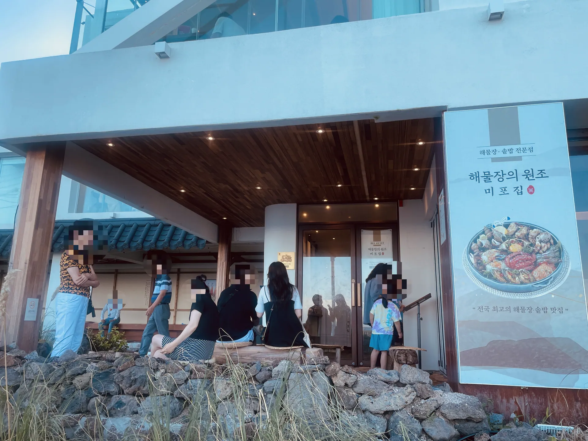 Entrance exterior of the exact Songjeong branch showing the restaurant name/sign and customers waiting outside. Visitors are incidental, not the photo subject; faces are pixelated by the source author. Kept as additional place context. Article published 2024-01-01T08:26:35+09:00; capture date unconfirmed.